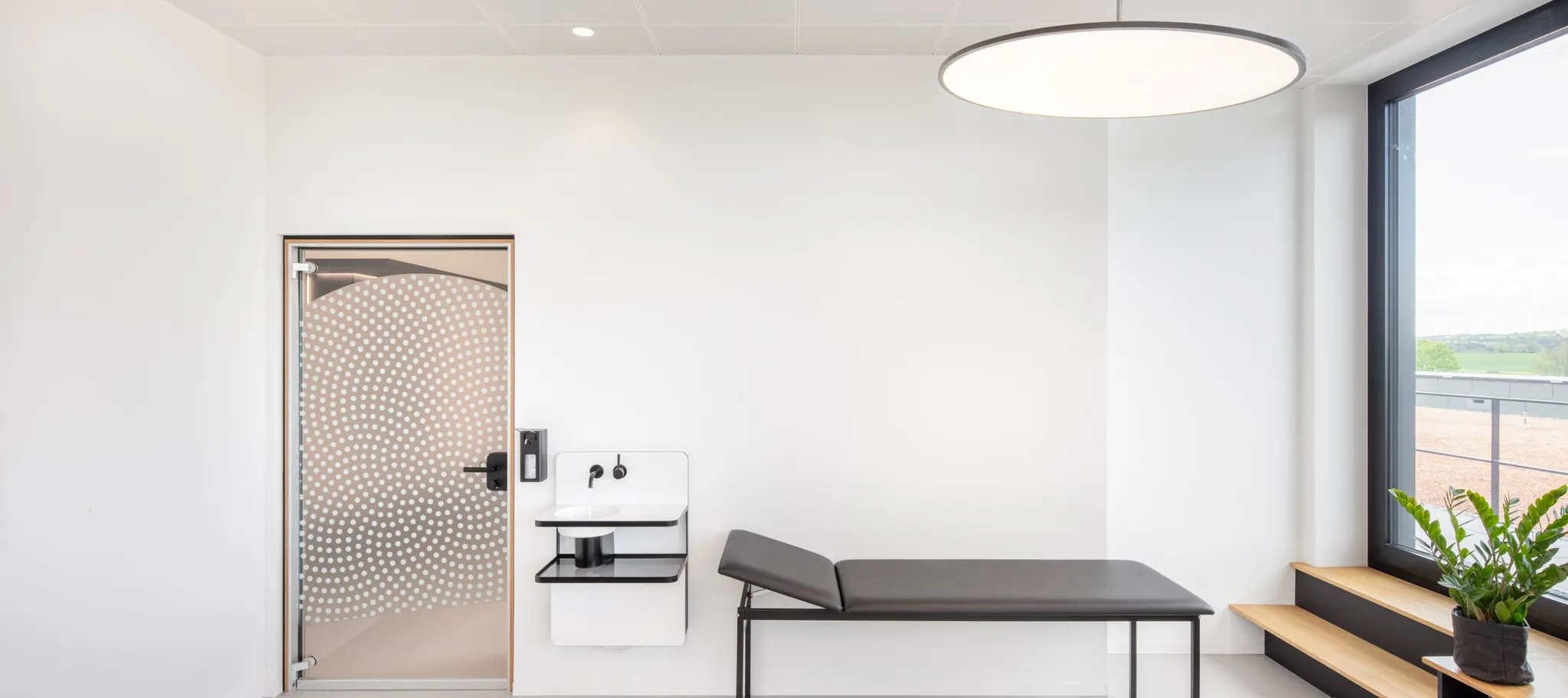 Modernes Behandlungszimmer mit großen Fenstern und einer medizinischen Liege auf grauem Betonboden